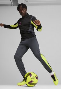 Atletische outfit bestaat uit een zwart en neon gele ritsjack, grijze broek en felgekleurde voetbalschoenen. Een levendige gele en roze voetbalsoccerbal ligt aan de voeten.