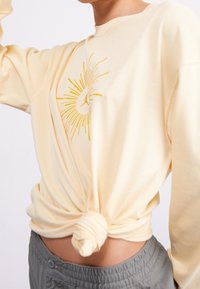 Person in einem hellgelben Sweatshirt, das um die Taille geknotet ist, mit einem gelben Sonnenstrahlenmuster, kombiniert mit grauen elasticisierten Hosen.