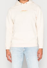 Kräms hoodie med en känguruficka, dragsko på huven och broderad "Santa Cruz"-logotyp i orange och gult. Har ribbade ärmslut och nederkant.