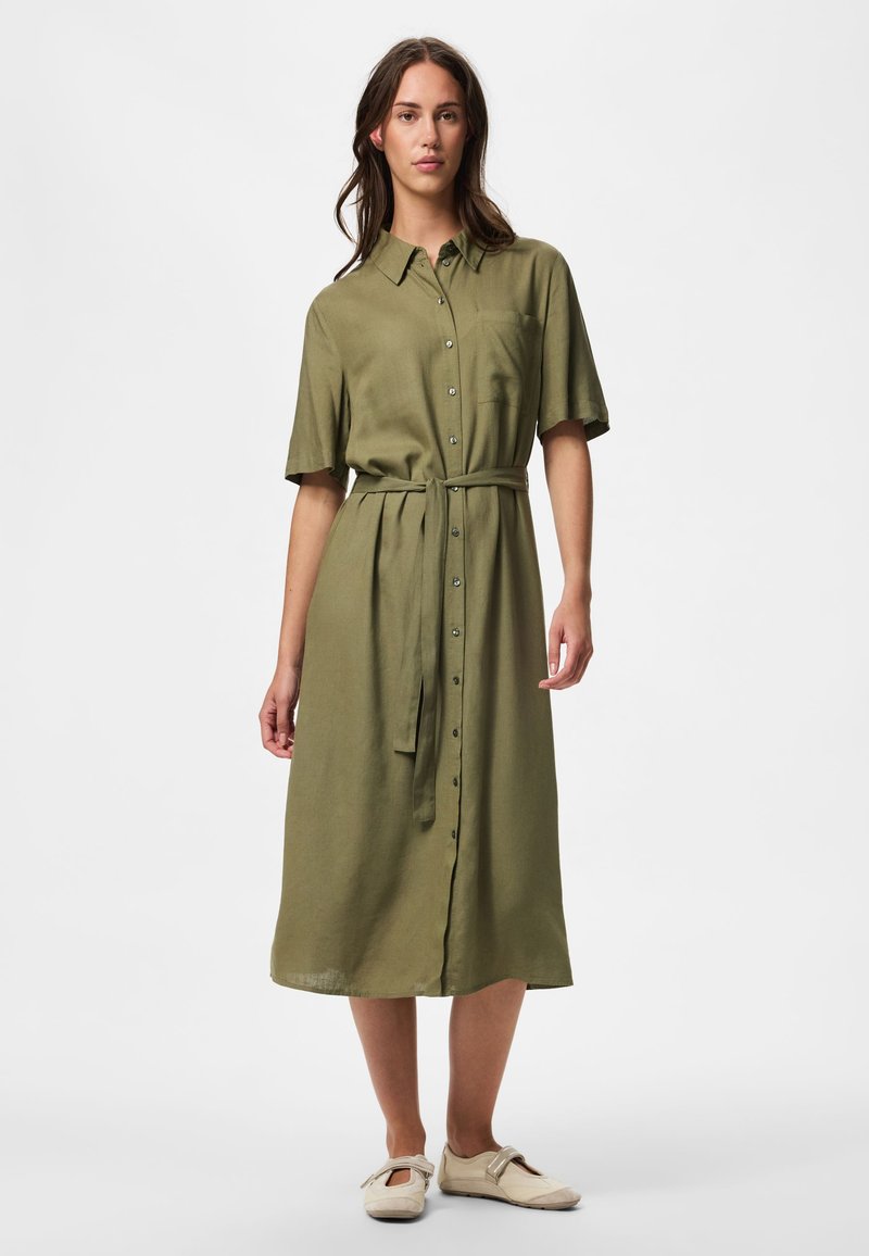 Femme portant une robe boutonnée olive mi-longue avec une ceinture à la taille et des chaussures Mary Jane beiges, se tenant devant un fond clair uni.
