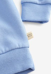 Felpa in cotone azzurro chiaro con polsini a costine. Presenta un piccolo etichetta di stoffa con la scritta "Little Bird". Texture morbida e design informale.