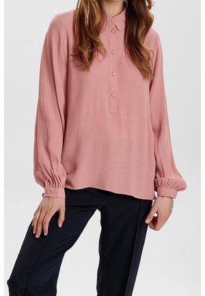 Femme portant un chemisier rose ample à manches longues avec des poignets boutonnés et un pantalon noir, debout devant un fond uni.