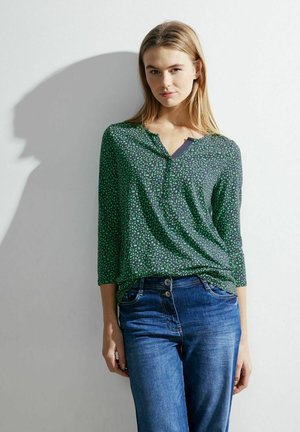 Blusa verde a fantasia con scollo a V e maniche a tre quarti, abbinata a jeans in denim blu. Tessuto morbido con un sottile disegno geometrico.
