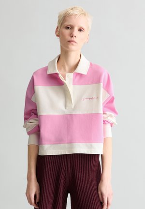 Persoană cu păr blond scurt, purtând un tricou rugby supradimensionat cu dungi roz și albe și pantaloni texturiți roșu închis, stând în fața unui fundal simplu.