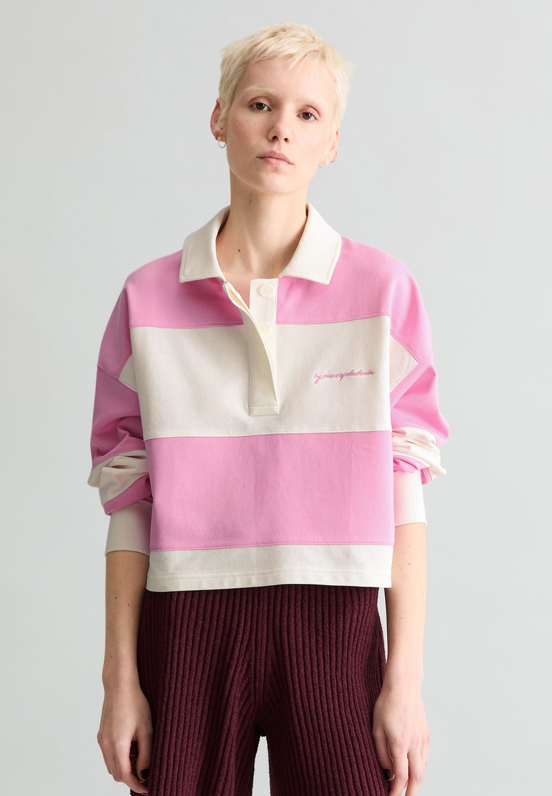 Person mit kurzen blonden Haaren, die ein übergroßes pink-weiß gestreiftes Rugbyhemd und strukturierte dunkelrote Hose trägt, vor einfachem Hintergrund steht.