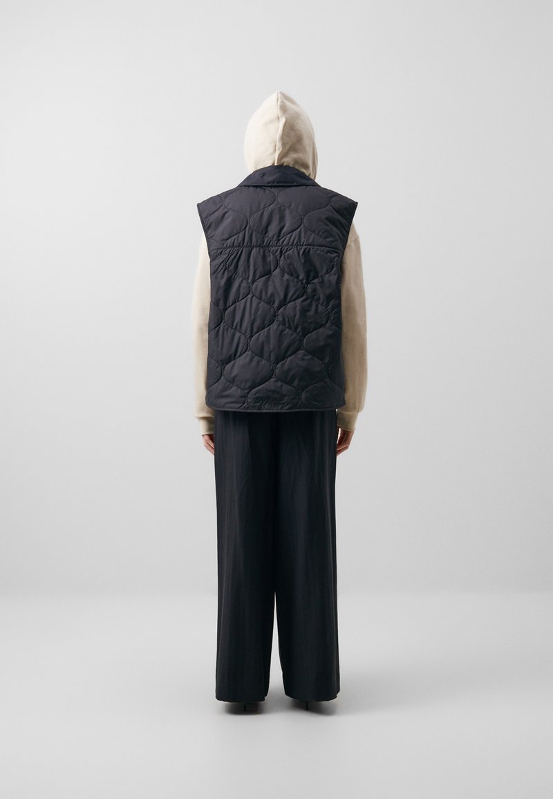 Gepolsterte schwarze Weste mit hohem Kragen und ärmellosem Design, getragen über einem hellen Kapuzenoberteil, kombiniert mit weiten schwarzen Hosen.
