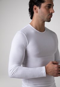 Camicia bianca a maniche lunghe realizzata in un tessuto liscio, con scollo rotondo e design aderente, caratterizzata da dettagli di cucitura discreti ai polsini.