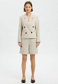 Lys beige linned blazer med dobbeltknaplukning, mørke knapper og matchende shorts, der snævrer ind ved lårene, båret med sorte mesh-flade sko.