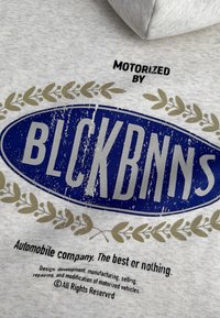 Grijze gebreide hoodie met een blauw ovale logo en vervaagde letters "BLCKBNNS" en gouden lauweren accenten eronder.
