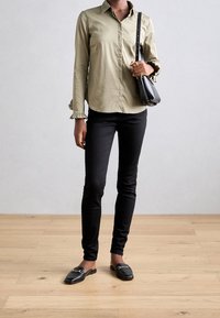Persona llevando una camisa de manga larga beige, pantalones ajustados negros, mocasines negros y sosteniendo un bolso negro en un suelo de madera.