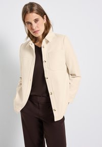 Kremowa kurtka koszulowa z kołnierzykiem, z zapięciem na guziki z przodu, dwiema kieszeniami na piersi i gładką fakturą, noszona na ciemno-brązowej bluzce i spodniach.