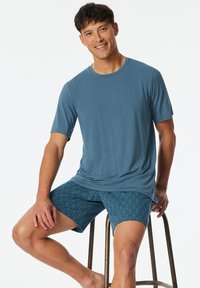 Kurzärmliges blaues Hemd, kombiniert mit türkisfarbenen Shorts mit geometrischem Muster; der Stoff wirkt glatt und leicht. Das Modell sitzt auf einem Metallhocker.