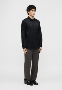 Άνδρας με σγουρά σκούρα μαλλιά, φορούν μαύρο πουκάμισο με κουμπιά, γκρι φαρδιά παντελόνια και μαύρα παπούτσια, στέκεται μπροστά σε λευκό φόντο.