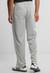 Lys grå treningsbukser med elastisk midje, med to baklommer og hvite side-striper. Glatt, blank stofftekstur.