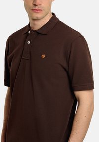 Camisa polo marrón de manga corta, con tapeta de tres botones y un pequeño logo naranja en el pecho. Hecha de una tela texturizada.