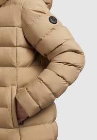 Beige Steppjacke mit gestepptem Design, versehen mit einem schwarzen Logopatch am Ärmel und einer Hand in der Tasche, glatte Stoffstruktur.