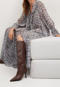 Nainen istuu valkoisella rahilla, päällään pitkä, rypytetty ja kuviollinen mekko sekä ruskeat polvipituiset korkokengät.