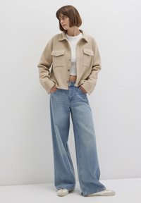Beige wool blend jacket with front pockets, paired with light blue wide-leg jeans. Model stands against a plain white background.