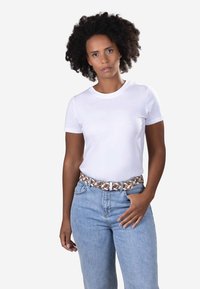 Vrouw met krullend haar, gekleed in een eenvoudig wit T-shirt, ingestopt in een lichtblauwe spijkerbroek met een gevlochten bruine en witte riem, één hand in de zak.