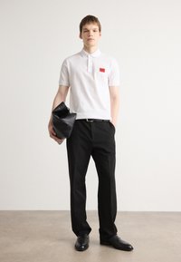 Hvid polo shirt med korte ærmer, rød logo patch; sorte bukser; holder en sort lædertaske; skinnende sorte sko; neutral baggrund.