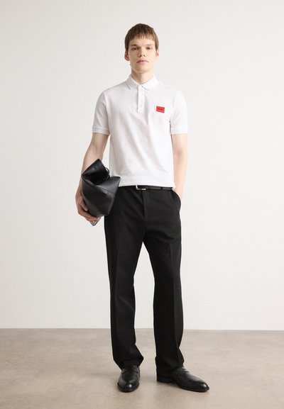 Polo blanc à manches courtes, écusson rouge ; pantalon noir ; portant un sac en cuir noir ; chaussures noires brillantes ; arrière-plan neutre.