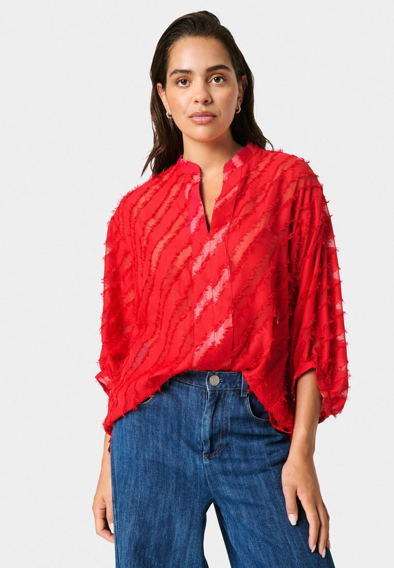 Blusa rossa semi-trasparente con strisce testurizzate verticali, colletto alla mandarino e vestibilità ampia, abbinata a jeans di denim blu.