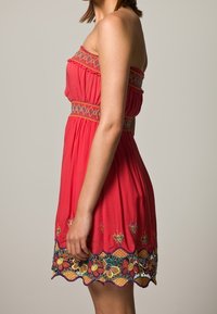 Vestido rojo sin mangas con patrones florales bordados y dobladillo en forma de ondas. El corpete y la cintura elásticos proporcionan un ajuste relajado. Tejido suave y liviano.