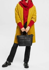 Schwarze Lederhandtasche mit zwei Reißverschlusstaschen, gehalten von einer Person, die einen gelben Mantel und einen roten Strickpullover, schwarze Hosen und glänzende Schuhe trägt.