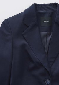 Blazer marine avec un col cranté, fermeture à un seul bouton et texture lisse. L'intérieur présente une doublure satinée. L'étiquette indique "un jour".