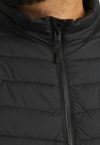 Schwarze gepolsterte Jacke mit hohem Kragen, voller Reißverschluss und einem subtilen Karomuster auf dem Stoff für zusätzliche Textur.