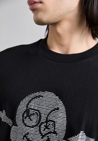 Svart bomull t-skjorte med et grafisk design i sølvprikker av to stiliserte ansikter på forsiden. Rund hals, kortermet.