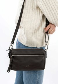 Borsa a tracolla in pelle marrone con tasca anteriore con zip, tracolla staccabile e dettagli in metallo argentato, appoggiata su un maglione chiaro.