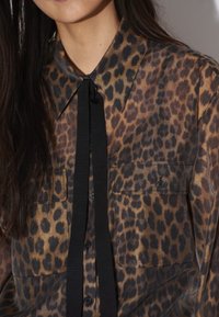 Doorschijnende blouse met luipaardprint en knopen, voorzien van een zwarte strik, met twee borstzakken en een klassieke kraag. Bruin en zwart kleurenpalet.