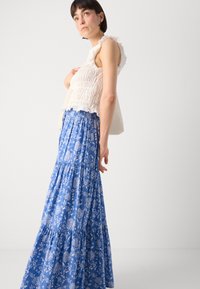 Weißes, gerafftes Crop-Top mit Rüschen, kombiniert mit einem langen, blauen Maxikleid mit floralen Mustern, das über gestufte Lagen und einen gerafften Taillenbund verfügt.