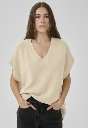 Jonge vrouw met lang bruin haar die een beige, mouwloze V-hals gebreide trui draagt en zwarte broek met trekkoord, handen in de zakken, neutrale achtergrond.