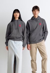 Woman and man wearing matching gray hoodies with red logo, woman in white skirt with slit, man in beige pants, standing side by side.