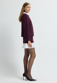 Blazer bordeaux avec col blanc, associé à une mini-jupe bordeaux avec un bas évasé blanc. Escarpins en cuir verni noir.