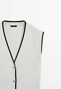 Cardigan blanc sans manches à col en V avec bordure noire et deux boutons sur le devant, présenté sur un fond blanc uni.