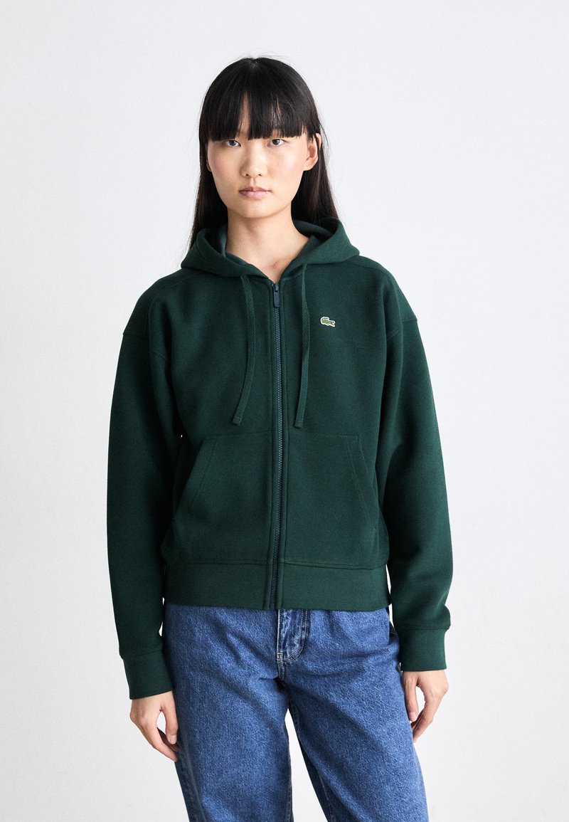 Grønn zip-up hettegenser i myk stoff, med en frontlomme og brodert logo på venstre bryst. Stilt med løse blåjeans.