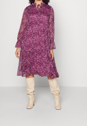 Vrouw die een paarse midi-jurk met lange mouwen en bloemenprint draagt, met doorschijnende mouwen en beige, slordige kniehoge laarzen, staand tegen een effen achtergrond.