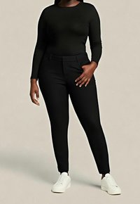 Personne portant un haut noir moulant à manches longues et un pantalon noir taille haute avec des baskets blanches, debout avec une main dans la poche.