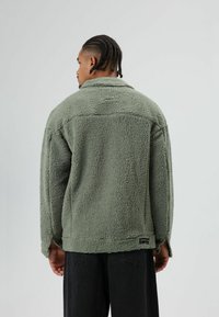 Green fleece jacket with a textured, soft exterior. It features a collar, long sleeves, and a small label at the hem. Worn over dark pants.