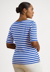 Woman with braided bun hairstyle wearing blue and white striped shirt and white pants, facing sideways against a plain background.