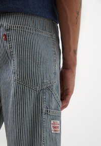 Pantalones de mezclilla a rayas azules y blancas con una etiqueta roja de Levi's, que cuentan con un bolsillo trasero con una etiqueta blanca que dice "trabajo por hacer".