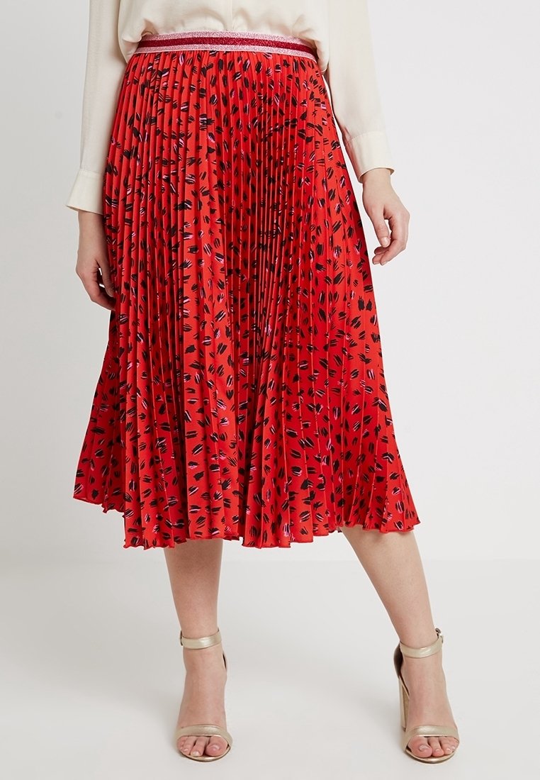 Femme portant une jupe midi plissée rouge avec des taches abstraites noires et roses et une ceinture rose métallique avec des sandales beiges à talons hauts.