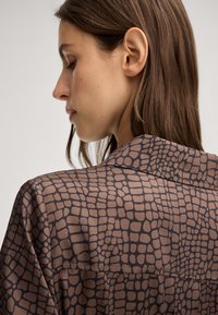 Blusa marrone a fantasia con una texture simile a quella di un rettile, con colletto e dettagli in bottone, mostrata da un'angolazione laterale su un modello.