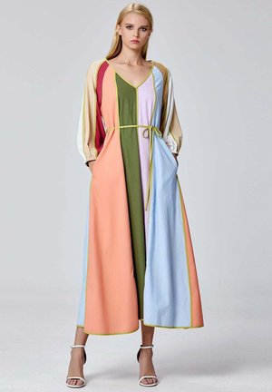 Femme portant une longue robe patchwork multicolore à manches longues et une fine ceinture jaune, debout les mains dans les poches.