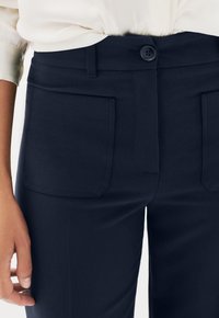 Gros plan sur un pantalon marine avec de grandes poches avant à rabat et une fermeture à bouton, porté avec une blouse beige clair rentrée.