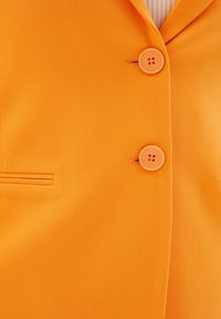 Pomarańczowa teksturowana marynarka z dwoma okrągłymi guzikami i pojedynczą kieszenią, z nowoczesnym kołnierzem.