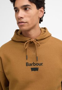 Καφέ φούτερ από βαμβάκι με κορδόνι, που διαθέτει το μαύρο λογότυπο "Barbour." και γραφικά από κάτω. Απαλή υφή και χαλαρός σχεδιασμός.